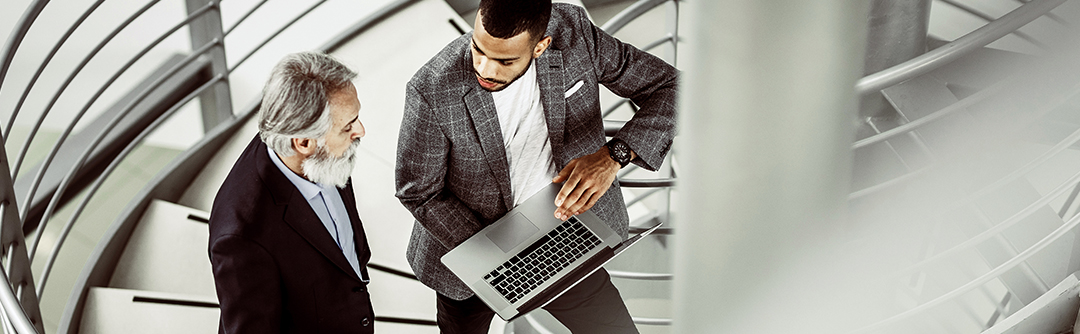 Two men standing on a staircase talking and looking at a laptop computer